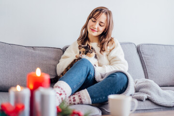 Woman in plaid with tea cup watching movie, TV, petting cat on sofa at home with christmas...