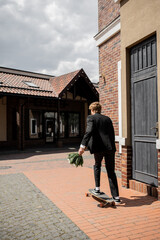 back view of groom in black suit holding wedding bouquet and  riding longboard on city street