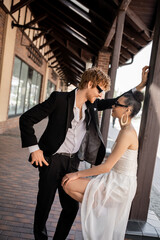 stylish redhead groom smiling near young african american bride on street, wedding in city