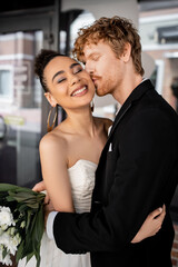 redhead groom in black suit embracing and kissing stylish african american bride on city street