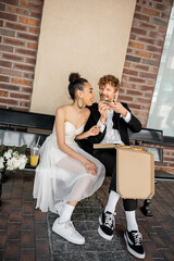 stylish and happy interracial newlyweds eating pizza near orange juice and flowers on bench in city