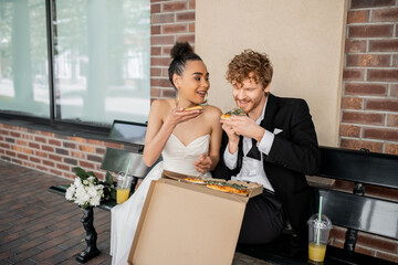 cheerful, elegant multiethnic newlyweds eating pizza near flowers and orange juice on bench in city