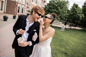 happy african american bride feeding stylish groom with french fries, wedding on city street