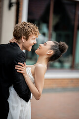 side view of young groom embracing overjoyed african american bride in city, outdoor celebration