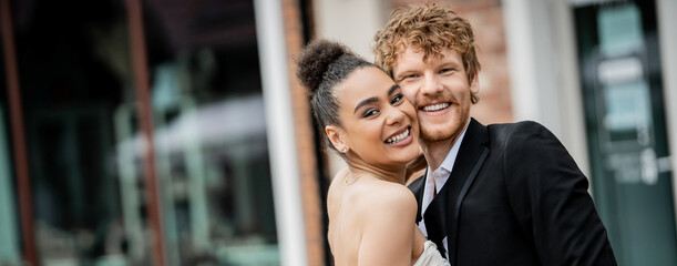 joyful interracial couple in wedding attire embracing and looking at camera on city street, banner