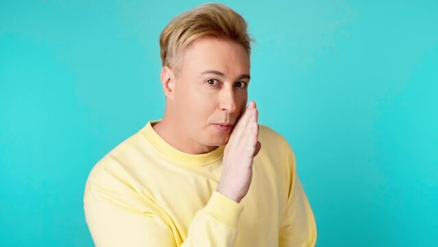 Secret, mystery and man keeps index finger on lips, makes silence gesture. Positive expression, confident smiling guy in yellow sweatshirt looks around in studio isolated on blue background.