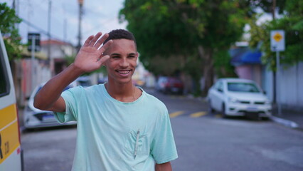 One cheerful young Brazilian man waving hello to neighbor walking in street. Joyful South American individual greeting friend with hand