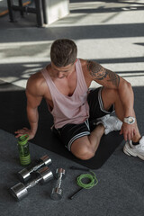 Handsome healthy muscular fitness guy with a hairstyle with a tattoo on his arm in sportswear sits on the floor with dumbbells, a skipping rope and a bottle of water in sport gym