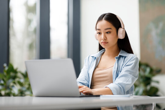 Asian Lady Student Wearing Headphones Taking Online Class On Laptop, Sitting At Table In University Classroom