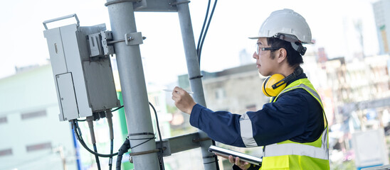 Building inspector man using digital tablet while checking telecommunication pole or telecom tower. Asian male technician inspecting and pointing at cellular phone network antenna on building rooftop - obrazy, fototapety, plakaty