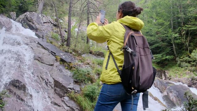 Young woman capturing waterfall images on a high mountain to share with friends using her mobile phone. Sharing on social media, family, nature, and healthy mountain life. Backpacker and blogger enjoy