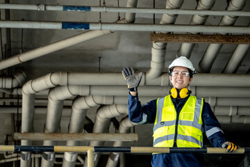 Fototapeta premium Asian male inspection engineer showing greeting gesture to say hello from upper floor. Cheerful man construction worker with reflective vest, protective goggles and ear muffs work at construction site