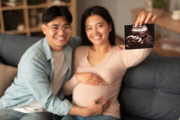 Cheerful pregnant korean spouses showing ultrasound photo sitting at home