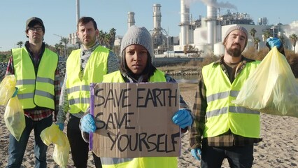 Group of volunteers looking at the camera seriously with garbage bags and a sign with the slogan save the planet save yourself.