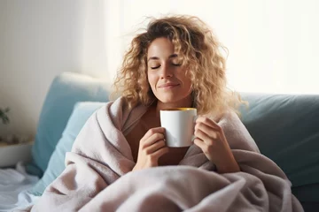 Fototapeten beautiful young blond dreamy woman sitting on bed wrapped in a blanket drinking tea or coffee © id512
