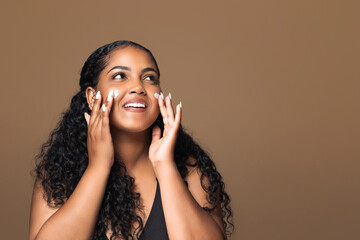 Female skincare. Happy latin body positive woman putting moisturizer cream on cheeks doing facial...