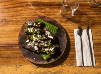 Top down view of traditional Mexican spicy dish with dark tortilla green herbs and sour cream served in wooden plate with metal dishes and white napkin