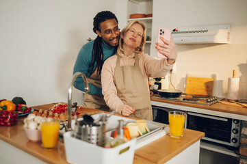 A happy multicultural couple in love is taking selfies in a kitchen while preparing lunch.