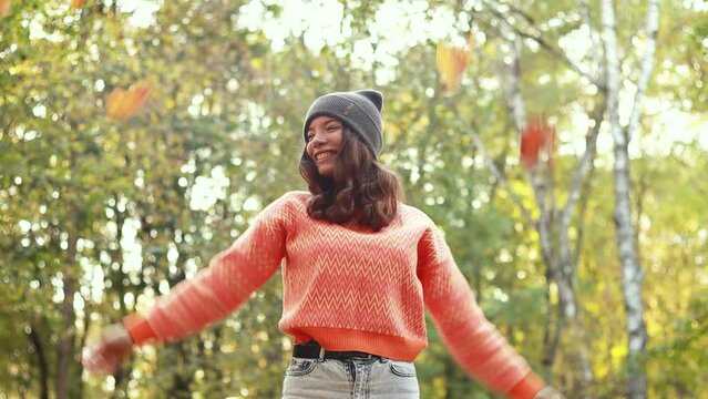 Slow motion footage. Caucasian woman walking outdoor, portrait of young lady in warm sunny autumn park season, fall, throwing yellow orange red leaves, dressed orange sweater