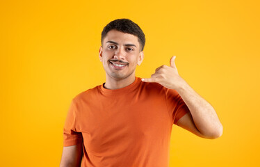 Cheerful brazilian man gesturing Call Me, holding hand like phone near ear and smiling, posing on yellow background