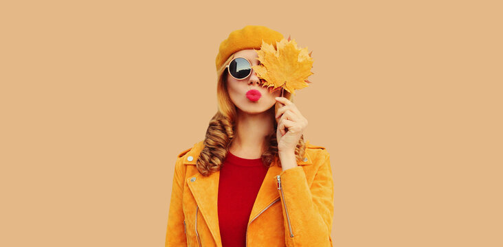 Autumn color style outfit, portrait of stylish beautiful young woman model with yellow maple leaves blowing her lips sends sweet kiss wearing orange french beret, sunglasses on brown studio background
