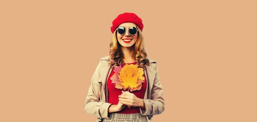 Autumn color style outfit, portrait of beautiful smiling young woman with yellow maple leaves wearing red french beret hat, jacket on brown studio background