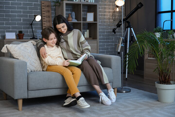 Fototapeta premium Little boy with his mother sitting on sofa and reading book at home late in evening