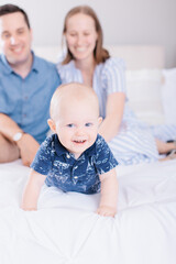 Fototapeta na wymiar One year old baby boy on bed with parents sitting behind him smiling