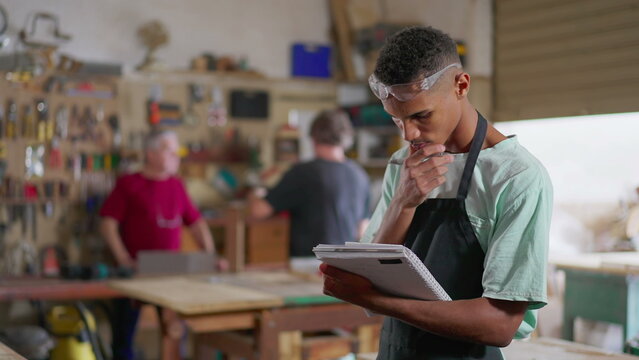 One young black apprentice of carpentry workshop taking notes on notepad, wearing apron and protective eye-wear. Staff employee engaged in learning business