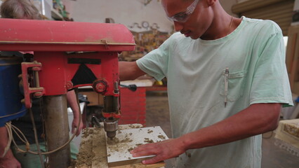 One young black Brazilian man engaged with his work using industrial machine drilling hole into wooden surface, person wearing protective eye-wear