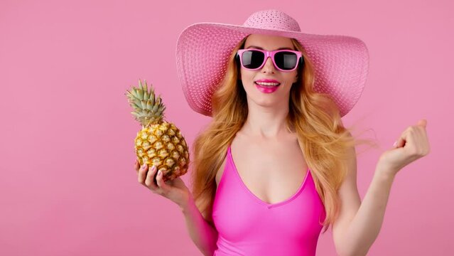 Fashionable woman dancing in swimsuit, total pink, tongue out, hat with pineapple, smiling enjoys summertime for spa, wellness. Playful glamor sexy lady dance, slim body, colorful trend on pink