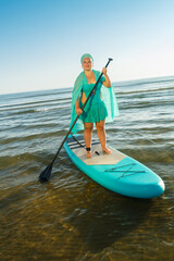 A Jewish woman in a turquoise swimsuit with a skirt and a scarf on her head on a SUP board swims to the seashore.