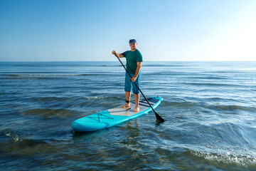 A man in shorts and a T-shirt stands on a SUP board with a paddle near the sea.