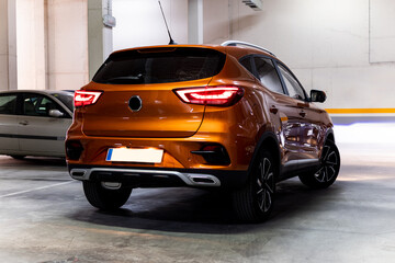 Back view of a modern orange SUV with its lights on is parked inside an underground car park. Car...