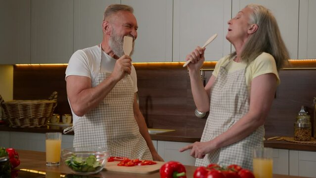 Mature gray wife and husband old couple grandparents sing song at home kitchen fun cooking lunch vegetable salad retired Caucasian man woman dance to music singing in kitchenware spoons food nutrition