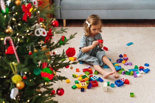 A little girl playing with colorful rainbow colored constructor pieces next to the christmas tree. Merry Christmas and Happy Holidays! Christmas gifts for children.