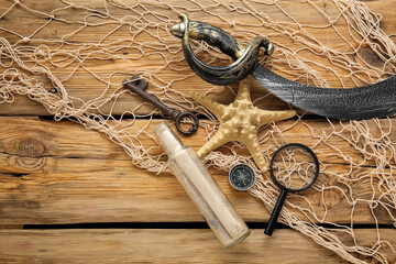 Pirate sword with scroll, magnifier, compass and net on brown wooden background