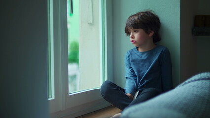 Bored child sitting indoors by apartment window looking out from second floor wanting to go outside, little boy with sad emotion - Powered by Adobe