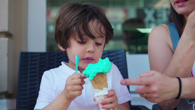 Child enjoying ice-cream cone at parlo shop during summer day. Mother brings cone closer to small boy's mouth to prevent from staining clothes
