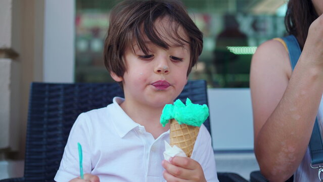 Child enjoying ice-cream cone at parlo shop during summer day. Mother brings cone closer to small boy's mouth to prevent from staining clothes