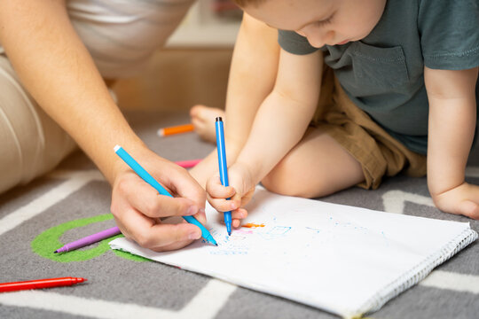 A beautiful little toddler boy of two or three years old with dad draws with markers in the album in the children's room at home sitting on the floor. Spending time with children. Selective focus