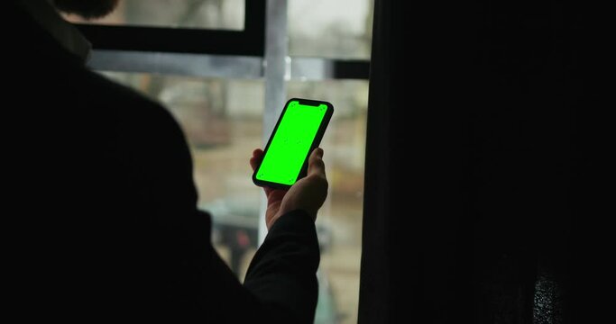 Male hand holding smart phone with green screen. Man using mobile phone while standing near window. Back view shot. Chroma key. Stay at home