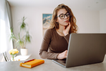 Young woman uses a laptop at home, looks at the screen, reads or writes at the table. Female works at home with a laptop. The concept of freelance, vacation, education.