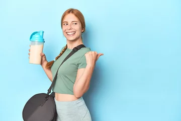 Zelfklevend Fotobehang Redhead holding protein shake in blue studio points with thumb finger away, laughing and carefree. © Asier