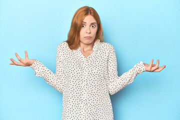 Redhead young woman on blue background doubting and shrugging shoulders in questioning gesture.