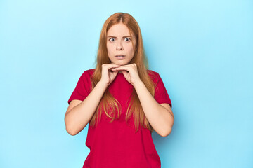 Redhead young woman on blue background praying for luck, amazed and opening mouth looking to front.