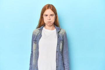 Redhead young woman on blue background blows cheeks, has tired expression. Facial expression...
