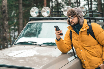 Young handsome male using mobile phone next to a jeep in the forest. Man wearing winter clothes...