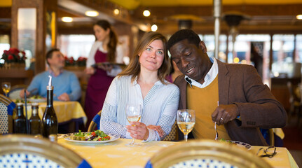 Cheerful adult biracial family couple enjoying delicious dinner in cozy restaurant. People sitting at served table, eating fresh vegetable salad and drinking wine
