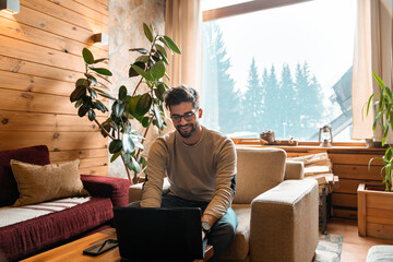 Handsome young adult using laptop while sitting indoors. Playful man surfing the internet in wooden...
